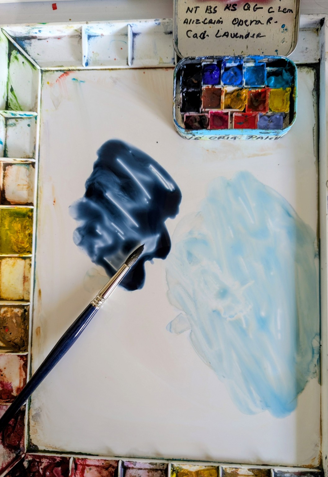 My open mint-tin palette sitting inside a large John Pike palette beside glossy dark and light blue washes and a pointed brush